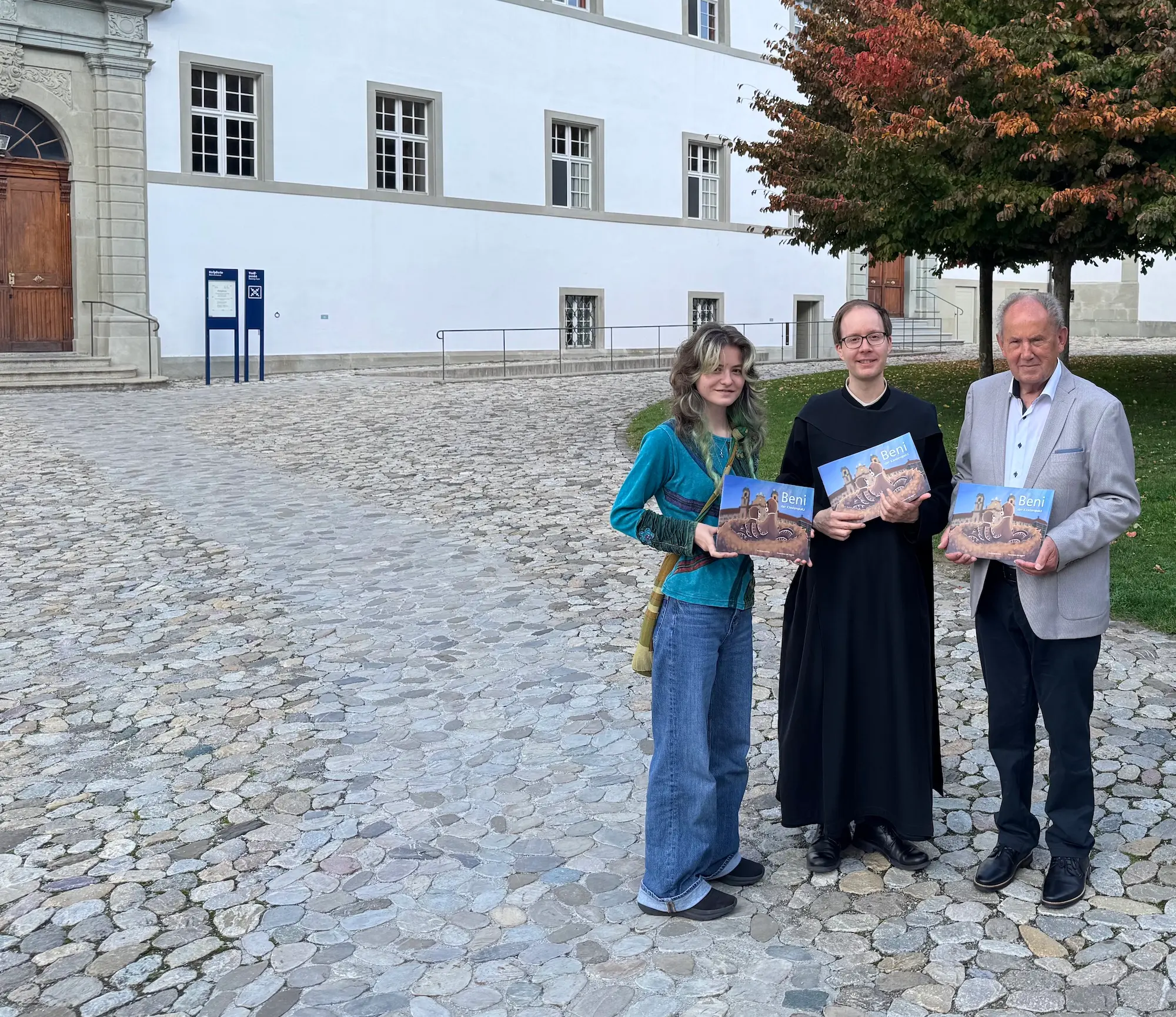 Das Foto, entstanden bei der Übergabe am 5. Oktober 2026, zeigt von links die Illustratorin Zoe Pertl, den Autor P. Thomas Fässler OSB und Verleger Josef Fink.